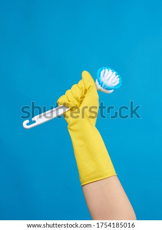 hand in a yellow rubber glove for cleaning a house holds plastic brush with a handle on a blue background