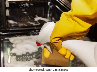 hand in a yellow rubber glove holds oven cleaner, cleaning the stove