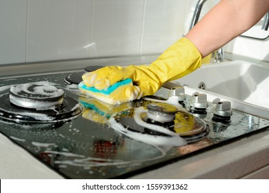 A hand in a yellow rubber glove washes a gas stove on a sunny day. Kitchen cleaning - Shutterstock ID 1559391362