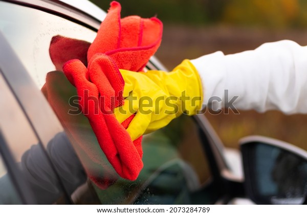 A hand in a yellow rubber glove\
wipes the glass of a car with a red microfiber rag from dirt on a\
warm autumn day. Wet cleaning. Selective focus.\
Close-up
