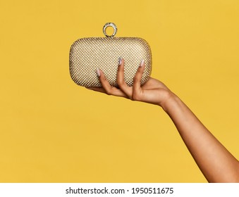 Hand Of A Young Black Woman Holding A Stylish Gold Metallic Pochette, Purse Or Small Handbag On Display Over A Yellow Studio Background With Copyspace