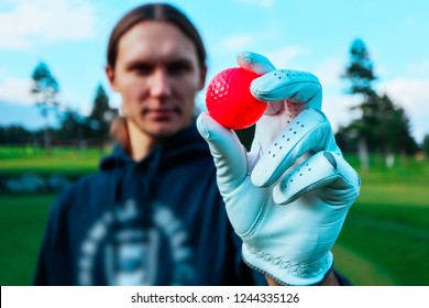 A Hand Of Young Golfer With A White Glove, Showing A Red Golf Ball, Close Up. Green Courses, Blue Sky And Mountains  On The Background.