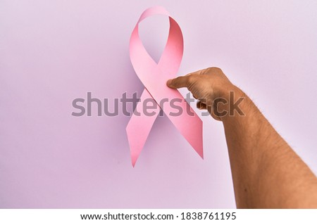 Hand of young hispanic man holding pink breast cancer ribbon over isolated background.