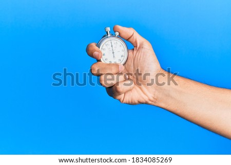Hand of young hispanic man using stopwatch over isolated blue background.