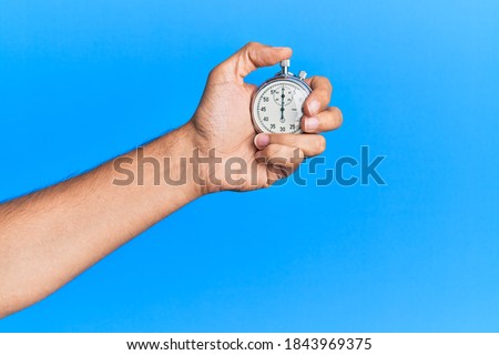 Hand of young hispanic man using stopwatch over isolated blue background.