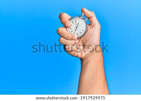 Hand of young hispanic man using stopwatch over isolated blue background.