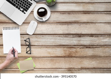 A Hand Of Young Man Is Going To Write On A Sketch Book. Wood Office Table With Laptop And Supplies, Top View With Copy Space.