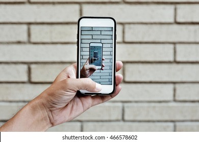 The Hand Of A Young Man Taking A Picture With Its Smartphone Of His Own Hand Taking A Picture With Its Smartphone Against A Brick Wall, As An Infinity Mirror Effect