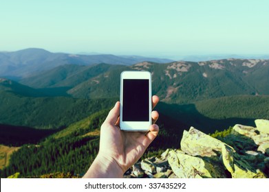 Hand Of Young Man, Which Is Standing On The Rock And Holding Smart Phone, With Blue Sky, Forests And Mountains On The Background, Close Up