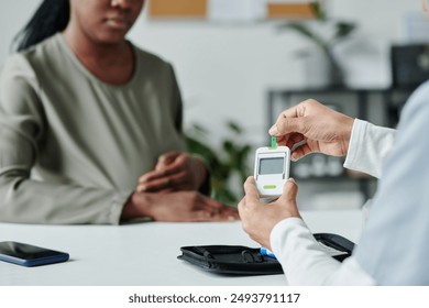 Hand of young unrecognizable female clinician with glucometer sitting by workplace in front of pregnant African American patient - Powered by Shutterstock