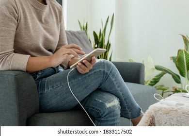 The Hand Of A Young Woman Holding A Smart Phone And A Modern Power Bank Charger. Put It At The Desk, Charging The Smartphone With A Spare Battery Charger, In Size That Sit Play Sofa.