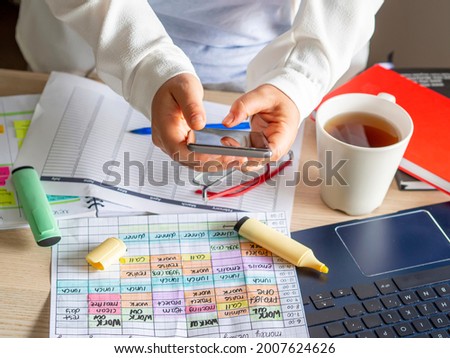 Hand of a young woman using smartphone to scan a planning calendar on chaotic desk in-home office. Work schedule and time Blocking technique for productivity. Desk with laptop, coffee and documents.