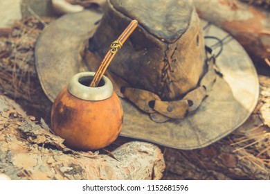 Handcrafted Artisan Yerba Mate Tea Calabash Gourd with Straw Leather Hat on Wood Logs in Forest. Travel Wanderlust Concept. Earthy Tones. Traditional Argentinian Latin American Brewing Cup
