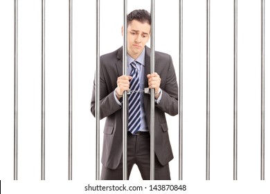 Handcuffed Businessman In Suit Posing In Jail And Holding Bars, Isolated On White Background