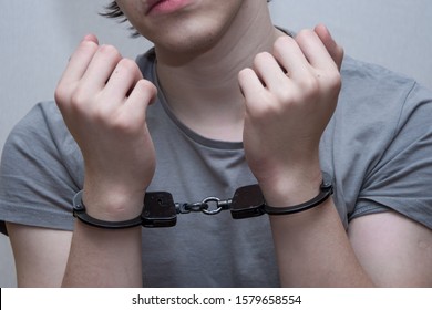 A Handcuffed Teenager Sits On A Grey Background. Juvenile Delinquent, Criminal Liability Of Minors. Members Of Youth Criminal Groups And Gangs.