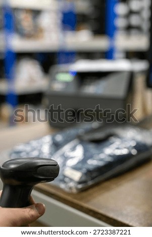 Handheld barcode scanner in use at a retail point-of-sale counter or warehouse, scanning a product with a label printer and storage racking in the background