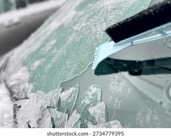 Handheld ice scraper clearing thick ice from car windshield during winter. Frosted glass, cold weather conditions, urban parking lot background, seasonal vehicle maintenance concept.