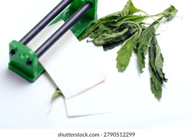 Handheld rolling tool with paper sheets and dried green herbal leaves for smoking on white background.