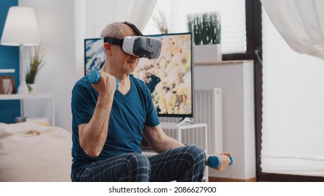 Handheld shot of elder man training with dumbbells and using vr glasses. Retired person lifting weights and wearing virtual reality headset to do physical exercise on fitness toning ball. - Powered by Shutterstock
