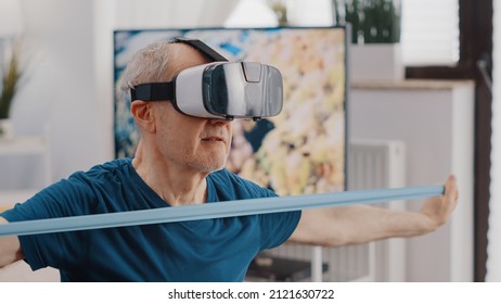 Handheld Shot Of Pensioner Pulling Resistance Band While Using Vr Glasses To Do Workout Training. Close Up Of Aged Man Doing Exercise With Elastic Belt And Wearing Virtual Reality Goggles