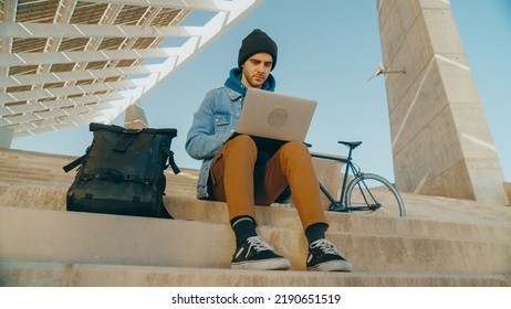 Handheld Shot Of Young Hipster Male Student Work On Laptop In Modern Urban Space In City. Cool Trendy Man. Commuter Freelancer With Bicycle And Backpack Work Remote In University