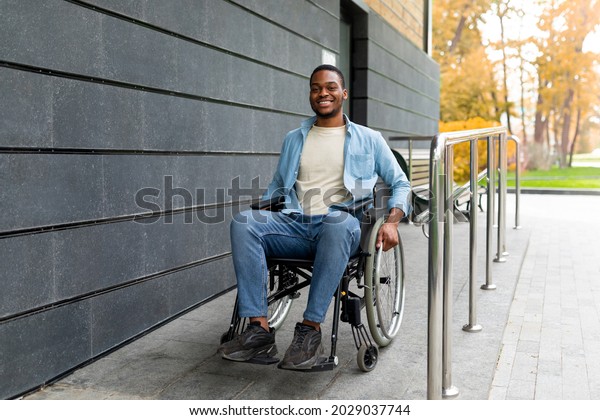 Handicapped accessible city concept. Positive\
impaired Afro man in wheelchair leaving building on ramp outdoors\
in autumn, full length. Joyful young black guy using disabled\
friendly facilities