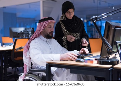 Handicapped Arabic businessman in tradition suit with female arabic assistent working in office. Disabled businessman in the wheelchair works in the office at the computer. 
