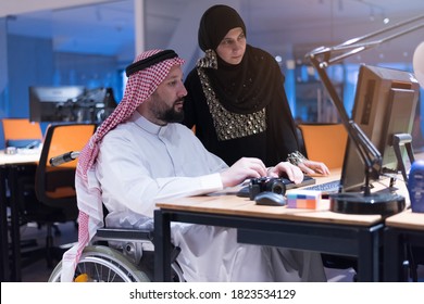 Handicapped Arabic businessman in tradition suit with female arabic assistent working in office. Disabled businessman in the wheelchair works in the office at the computer.