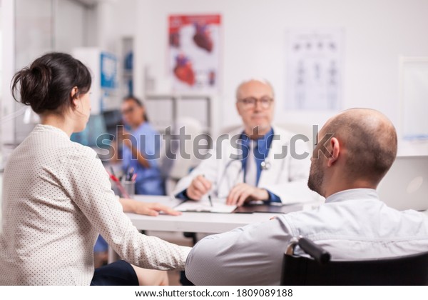 Handicapped\
man in wheelchair after car accident with wife at hospital for\
periodic check up with doctor. Senior medic explaining illness to\
disabled patient. Nurse holding x-ray\
image.