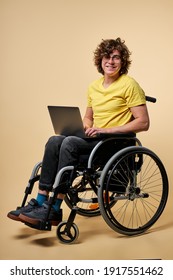 behinderte Person in zwangloser Kleidung sitzend mit Laptop auf Rollstuhl, einzeln auf beigem Studiohintergrund. gut aussehende, hässliche männliche Person, die gerne online arbeitet, Schreibnachricht auf der Tastatur