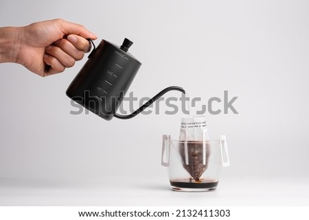 Handing holding black steel long spout drop kettle pouring hot water into a drip coffee bag in transparent cup. Brewing coffee trend at home. Isolated white background