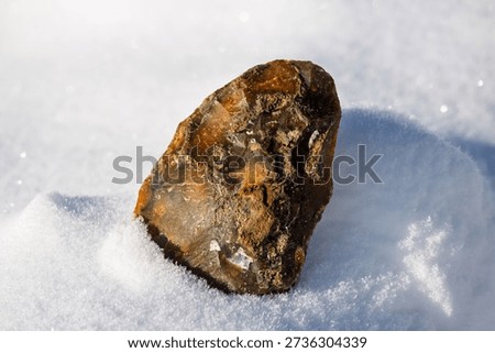 Hand-knapped primitive flint tool replica rests on sparkling pristine snow under bright sun. Ancient Paleolithic craftsmanship artifact