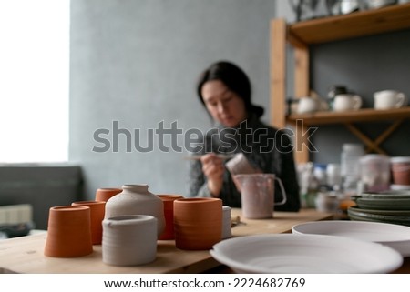 Handmade ceramic craft ware - empty cups, plates and bowls with female ceramics maker working on background. Woman making pottery item at own art studio.