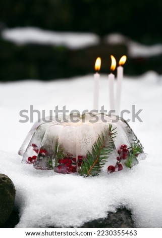Handmade Christmas lantern in shape of cake made of ice, red rose hips berries, holly berry branches and thuja twigs with burning candles in snow. Garden outdoor decoration concept. 