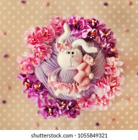 Handmade knitted toy. Easter Bunny in a pink skirt and a bow on his head holds a plush pink Teddy bear in his paw against the background of purple and pink flowers. Beige dots background. - Powered by Shutterstock