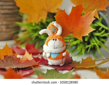 Handmade knitted toy. Easter Bunny in orange sweater and hat with pompom on the background of bright autumn maple leaves - Powered by Shutterstock