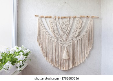 Handmade macrame. 100% cotton wall decoration with wooden stick hanging on a white wall. Female hobby. ECO friendly modern knitting DIY natural decoration concept in the interior