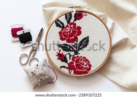 Handmade red and black cross-stitch floral ornament of red roses on white linen fabric in wooden hoop. Needlework and DIY concept. Embroidery as a hobby. Top view, close-up, flat lay