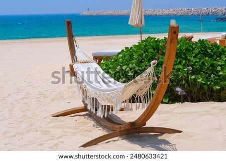  Handmade white hammock-swing on solid wooden base close up at tropical paradise ocean beach in bright sunny summer day. Summer beach vacation or luxury romantic holiday background. 