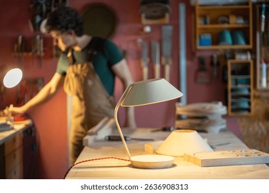 Handmade wooden lamp with unique design stands lit on table in workshop. Carpenter making notes while working in garage shop background, custom creation, warm lighting business, artisan workspace  - Powered by Shutterstock - Get 15% off with code: PIKWIZARD15
