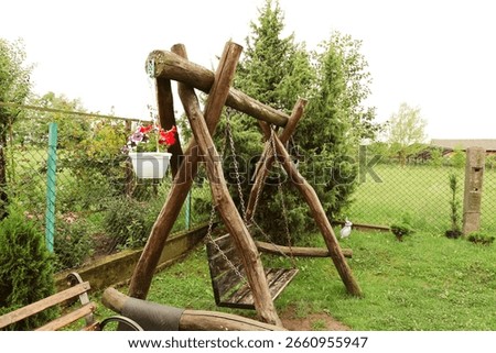 A handmade wooden swing made of solid wood stands in a cozy garden. A pot with bright flowers hangs nearby, creating an atmosphere of peaceful relaxation and family leisure in the fresh air.
