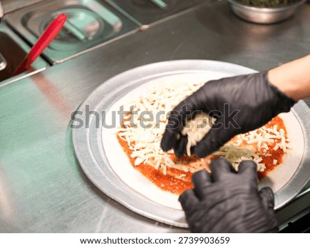 Hands adding shredded cheese to a pizza base with tomato sauce on a metal tray, pizza preparation in kitchen.