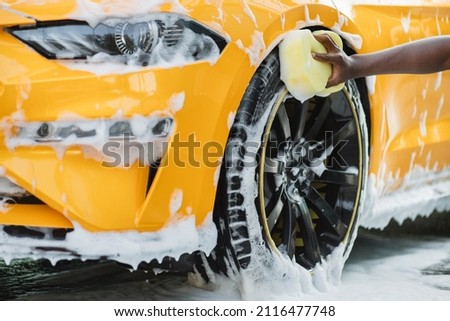 Hands of African man holding yellow sponge, washing car wheel with foam. Cleaning of modern rims of luxury yellow car at self car wash service outdoors.