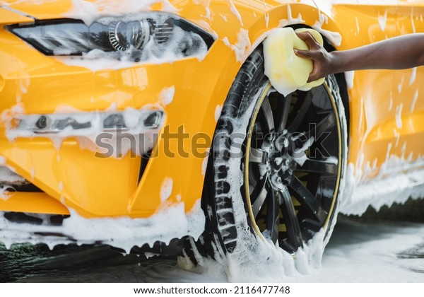 Hands of African man holding yellow\
sponge, washing car wheel with foam. Cleaning of modern rims of\
luxury yellow car at self car wash service\
outdoors.