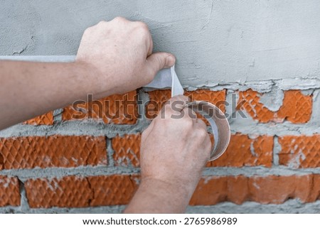 Hands apply tape to brick wall. Fingers grip tape, pulling it taut. Surface shows cracked mortar, exposed brick