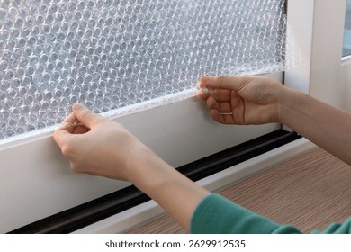 Hands applying bubble wrap to a window for insulation in a cozy indoor setting during the winter season