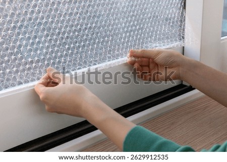 Hands applying bubble wrap to a window for insulation in a cozy indoor setting during the winter season