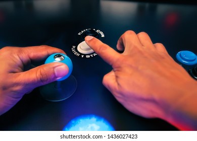 The hands of an arcade player reaching for the player one start button and holding a top fire four way joystick.