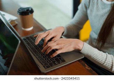 The hands of an Asian businesswoman in gray long-sleeved sweater owns coffee shop. Typing coffee product revenue summary into laptop. Next to disposable paper coffee cups small business coffee shop - Powered by Shutterstock