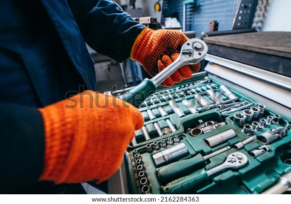 Hands of\
auto mechanic in yellow work gloves holding wrenches above a set of\
tools from wrenches and heads for unscrewing nuts and bolts in a\
special cabinet for repair in a car\
service.